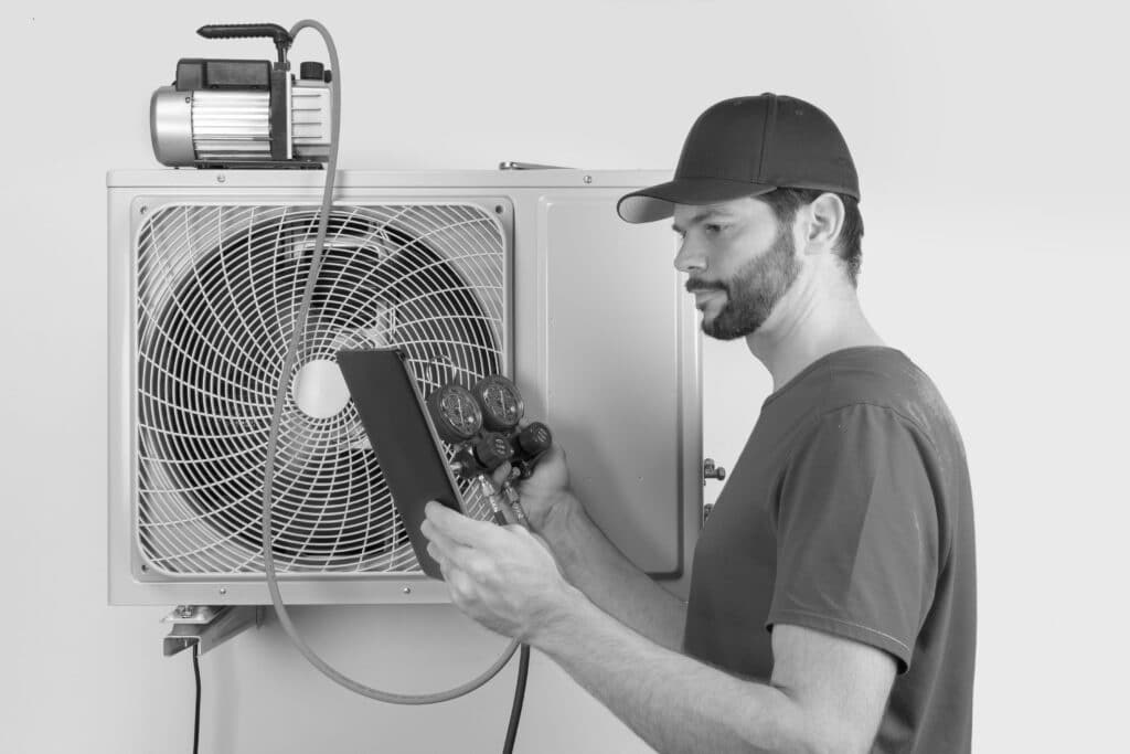 Installation service fix repair maintenance of an air conditioner outdoor unit, by cryogenist technican worker evacuate the system with vacuum pump and manifold gauges tablet in blue shirt and baseball cap